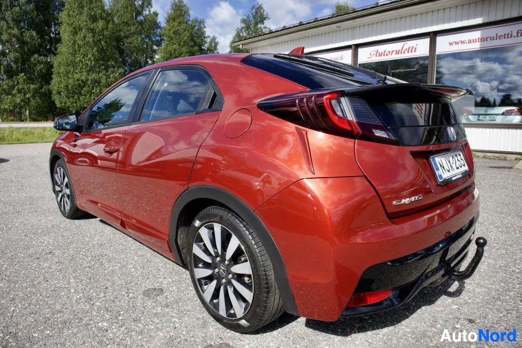 Takaviisto: Oranssinpunainen henkilöauto takaa oikealta kuvattuna, pysäköitynä ulos. Auton takavalot ovat näkyvissä, ja siinä on vetokoukku. Taustalla näkyy rakennus, jossa