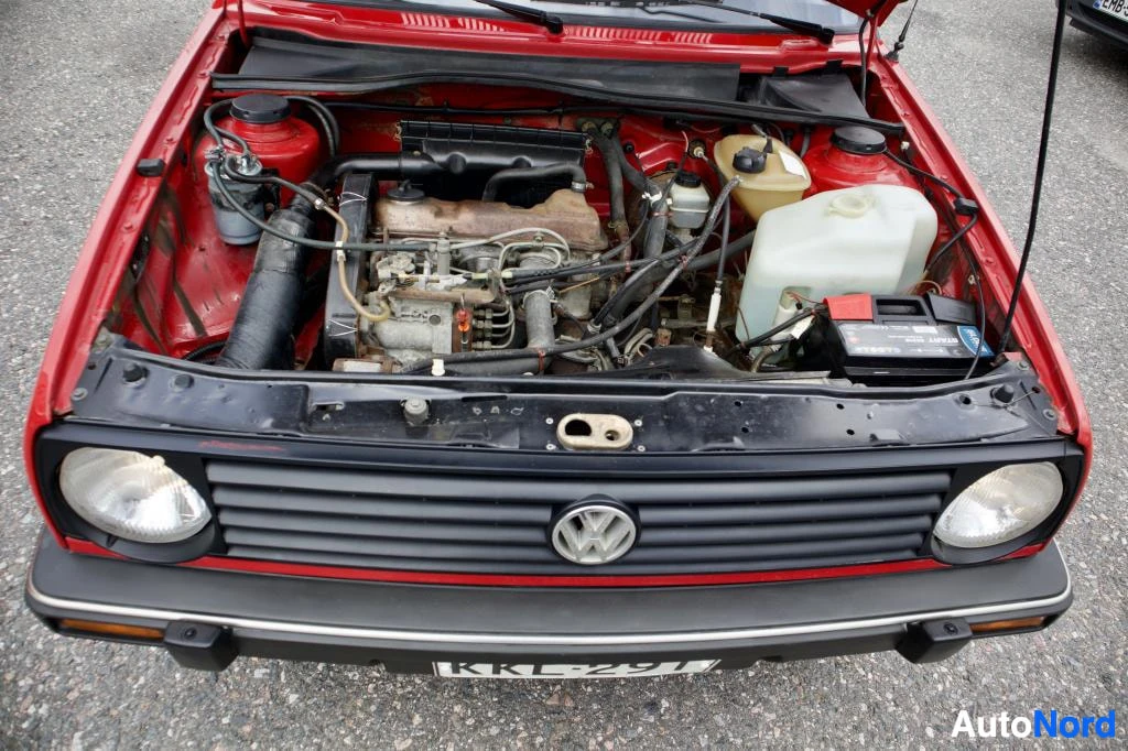 Moottoritila: Kuva auton moottoritilasta edestä päin. Konepelti on auki. Auton keula on punainen. Volkswagen-logo ja rekisterikilpi "" näkyvät. Akku ja pesunestesäiliö