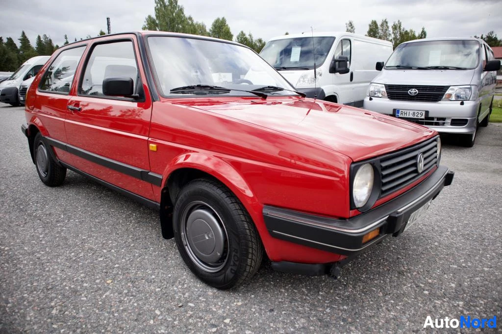Etuviisto, nokka oikealle: Punainen henkilöauto pysäköitynä. Ajoneuvon etuosa ja oikea kylki ovat näkyvissä. Autossa on mustat puskurit ja mustat sivupeilit.