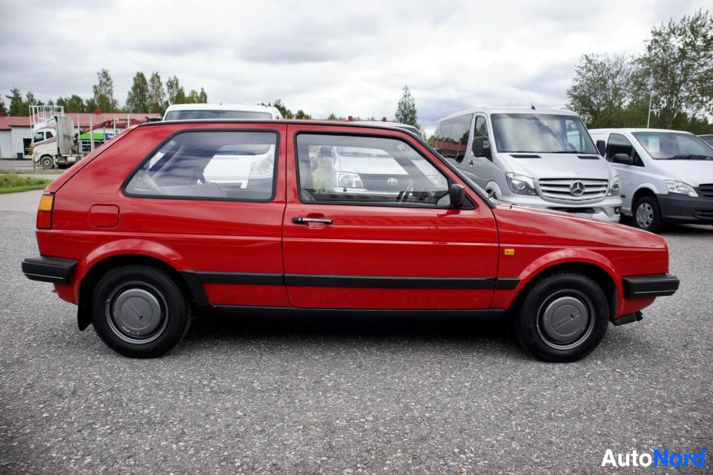 Sivulta: Punainen henkilöauto pysäköitynä. Ajoneuvon vasen kylki on näkyvissä. Autossa on kaksi ovea ja mustat puskurit.