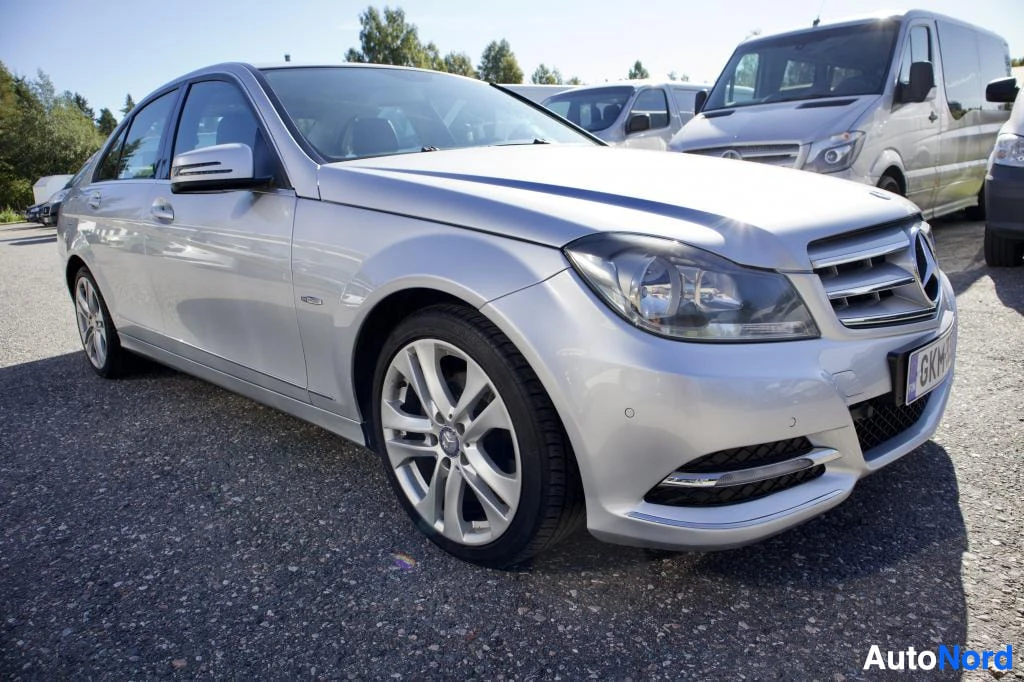 Etuviisto, nokka oikealle: Hopeanvärinen Mercedes-Benz C-sarjan sedan pysäköitynä ulos aurinkoisena päivänä. Auton keula ja vasen kylki näkyvät etuviistosta. Edessä on näkyvillä re
