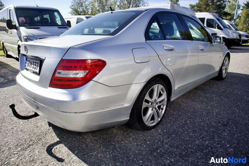 Takaviisto: Hopeanvärinen Mercedes-Benz C-sarjan sedanin takaosan kolmen neljänneksen kuva vasemmalta puolelta, vetokoukku näkyvissä. Auton takarekisterikilvessä on teksti