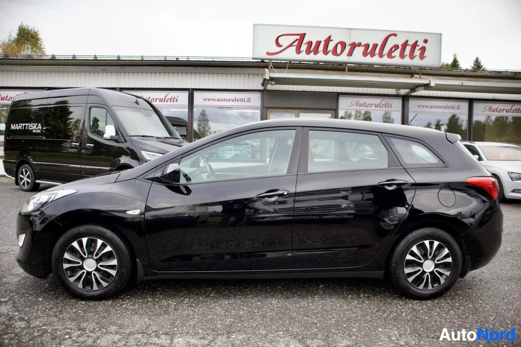 Sivulta: Musta Hyundai i30 -farmariauto kuvattuna suoraan sivulta ulkona pysäköitynä rakennuksen edessä. Takana näkyy musta pakettiauto.