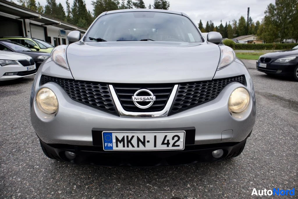 Edestä: Hopeanvärinen Nissan Juke edestä kuvattuna parkkipaikalla. Ajovalot ovat päällä. Etupuskurissa on rekisterikilpi.
