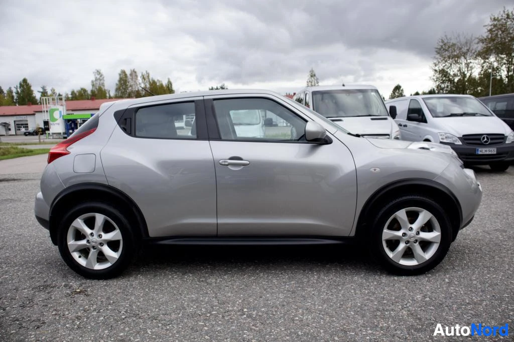 Sivulta: Hopeanvärinen henkilöauto oikealla sivulla parkkipaikalla, taustalla rakennuksia ja pilvinen taivas. Autossa on viisipuolaiset kevytmetallivanteet.