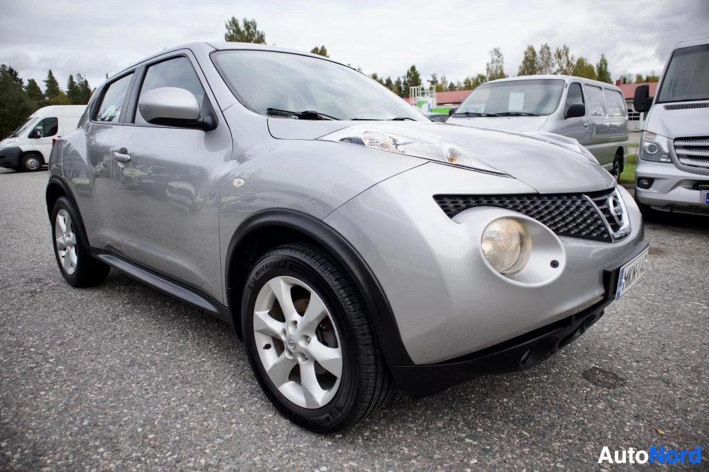 Etuviisto, nokka oikealle: Hopeanvärinen Nissan Juke etuoikealta kuvattuna parkkipaikalla muiden autojen vieressä. Ajovalot ovat päällä ja vanteet ovat hopeanväriset monipuolavante