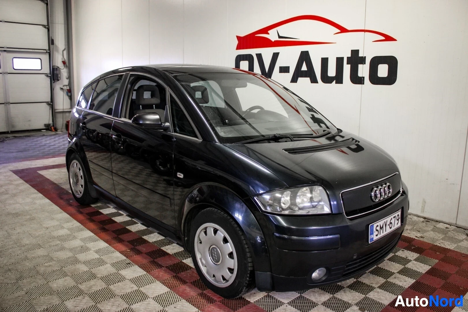 Etuviisto, nokka oikealle: Musta Audi A2 edestä ja sivulta kuvattuna sisätiloissa. Autossa on rekisterikilpi ja vanteet ovat harmaat metallikapselit