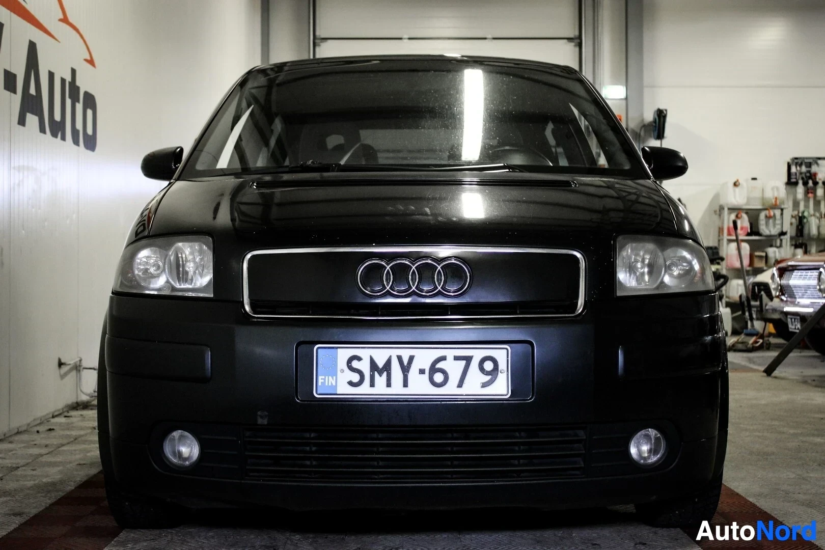Edestä: Musta Audi A2 edestä kuvattuna sisätiloissa. Autossa on rekisterikilpi