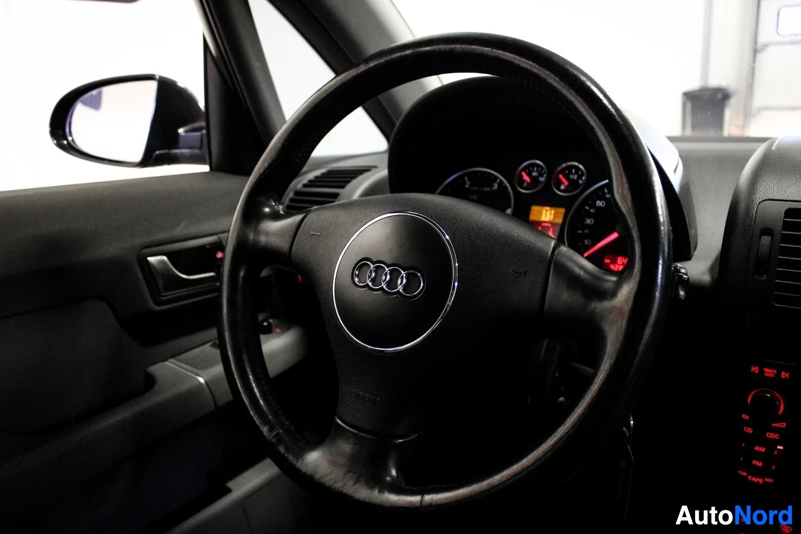 Ohjaamo: Kuva auton sisätiloista kuljettajan paikalta, keskittyen ohjauspyörään ja mittaristoon. Ohjauspyörässä on Audi-logo keskellä. Taustapeili näkyy kuvan vasemmassa
