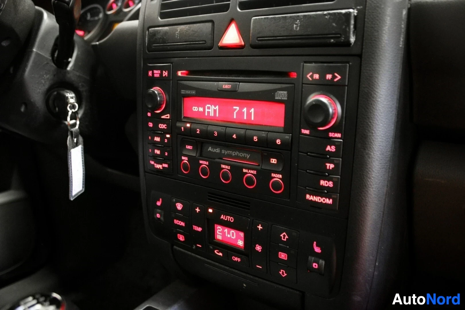 Ohjaamo: Lähikuva auton keskikonsolista, jossa on Audi Symphony -radio/CD-soitin ja ilmastoinnin säätimet, joissa on punainen taustavalo. Käsijarru ja auton avaimet näky