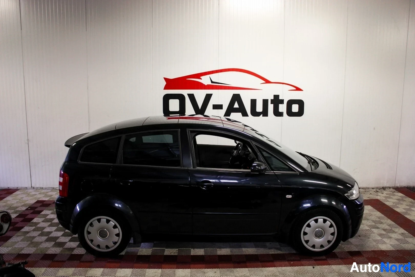 Sivulta: Musta henkilöauto kuvattuna suoraan oikealta sivulta. Auto on parkissa hallissa, jonka seinällä on OV-Auto-logo. Autossa on hopeanväriset vanteet.