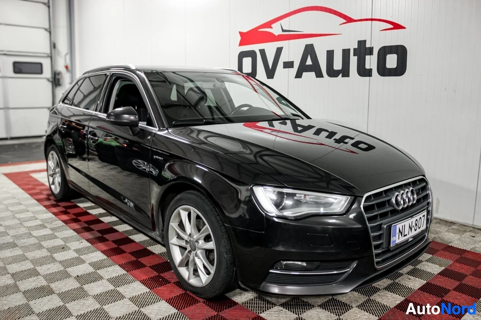 Etuviisto, nokka vasemmalle: Musta Audi A3 Sportback pysäköitynä viistoittain sisätiloissa, etuosan vasen puoli ja kylki näkyvissä. Eturengas on kääntynyt hieman vasemmalle. Auton