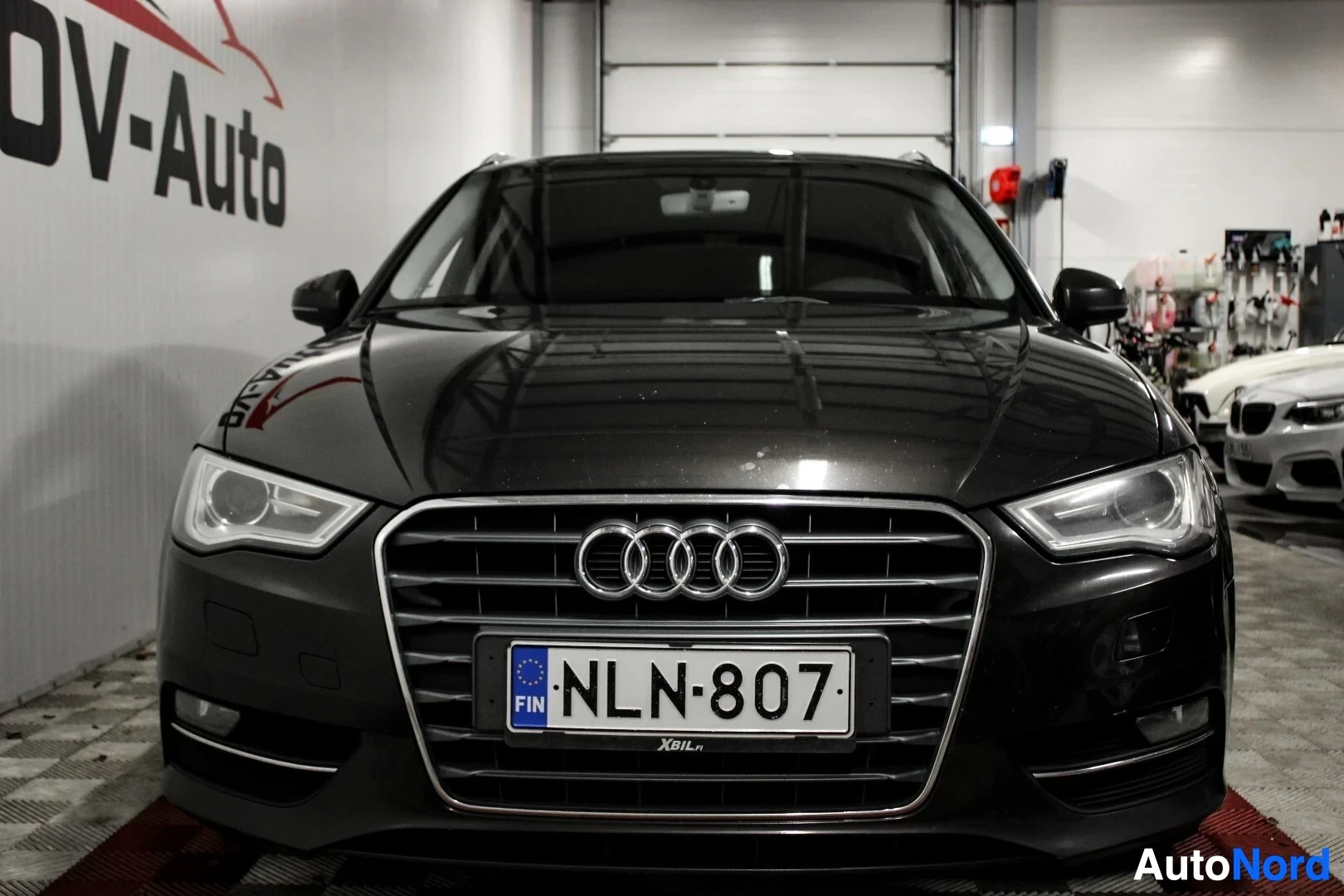Edestä: Mustan Audi A3 Sportbackin etuosa on kuvattu suoraan edestä. Etusäleikkö, ajovalot ja rekisterikilpi "" näkyvät selvästi. Taustalla on valkoinen seinä, j