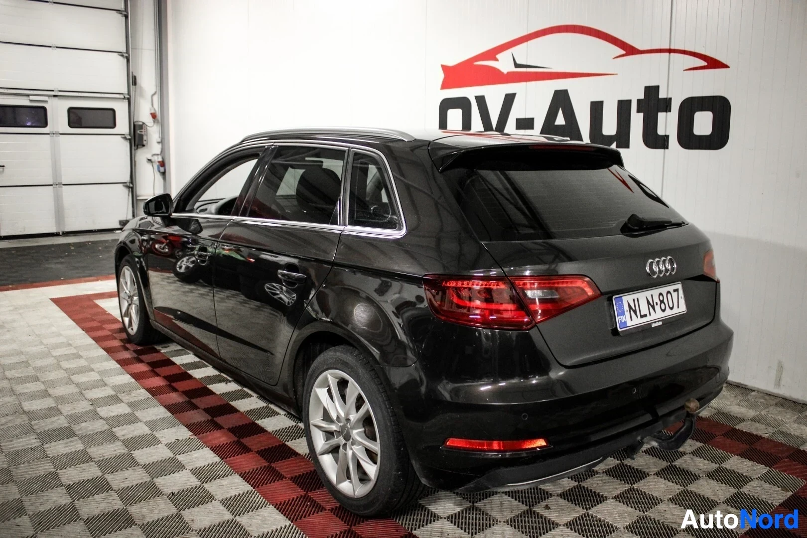 Takaviisto: Musta Audi A3 Sportback on pysäköity viistoittain sisätiloissa, auton takapää ja vasen kylki näkyvissä. Rekisterikilvessä lukee "". Takana näkyy vetokouk