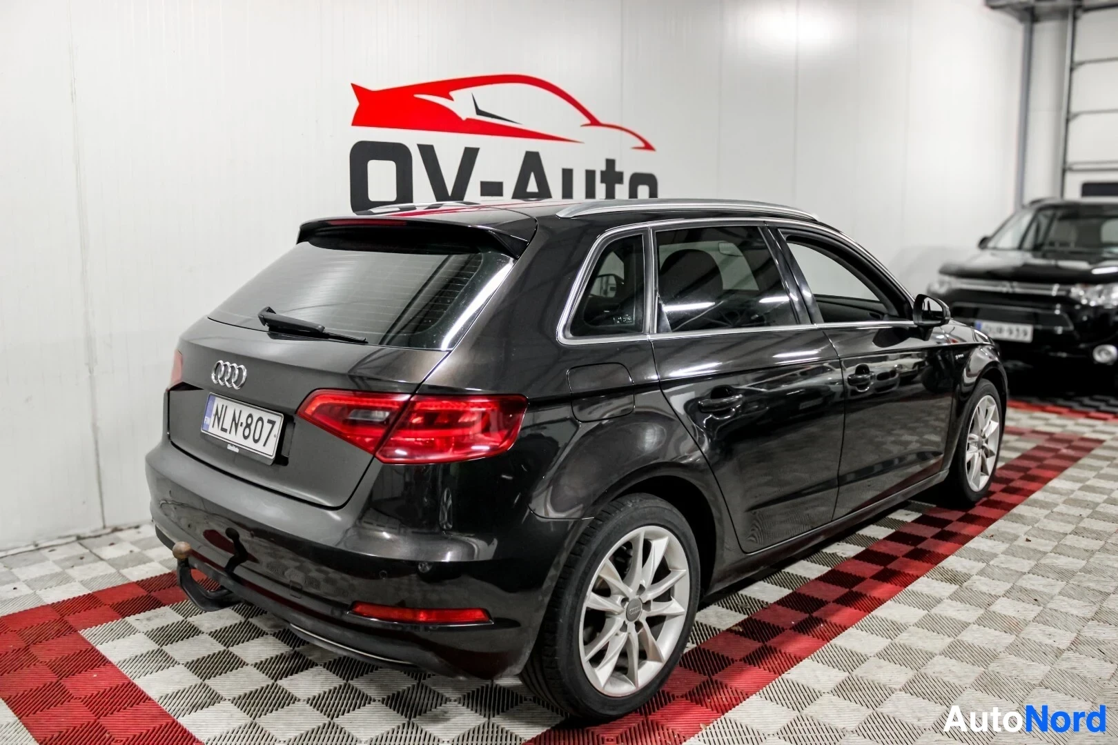 Takaviisto: Musta Audi A3 Sportback auton takaa ja oikealta sivulta kuvattuna. Autossa on hopeiset kevytmetallivanteet ja vetokoukku. Rekisterikilpi näkyy auton tak