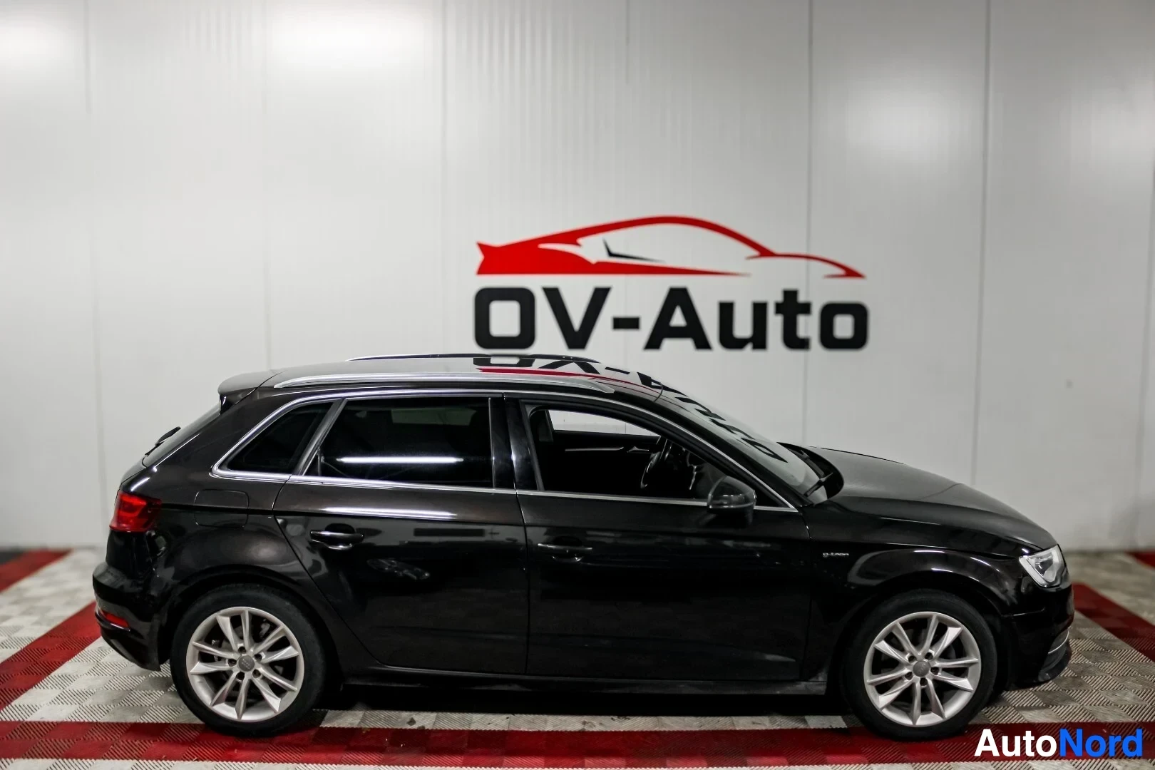 Sivulta: Musta Audi A3 Sportback auton vasemmalta sivulta kuvattuna. Autossa on hopeiset kevytmetallivanteet ja kattokiskot. Taustalla on valkoinen seinä, jossa on OV-Au