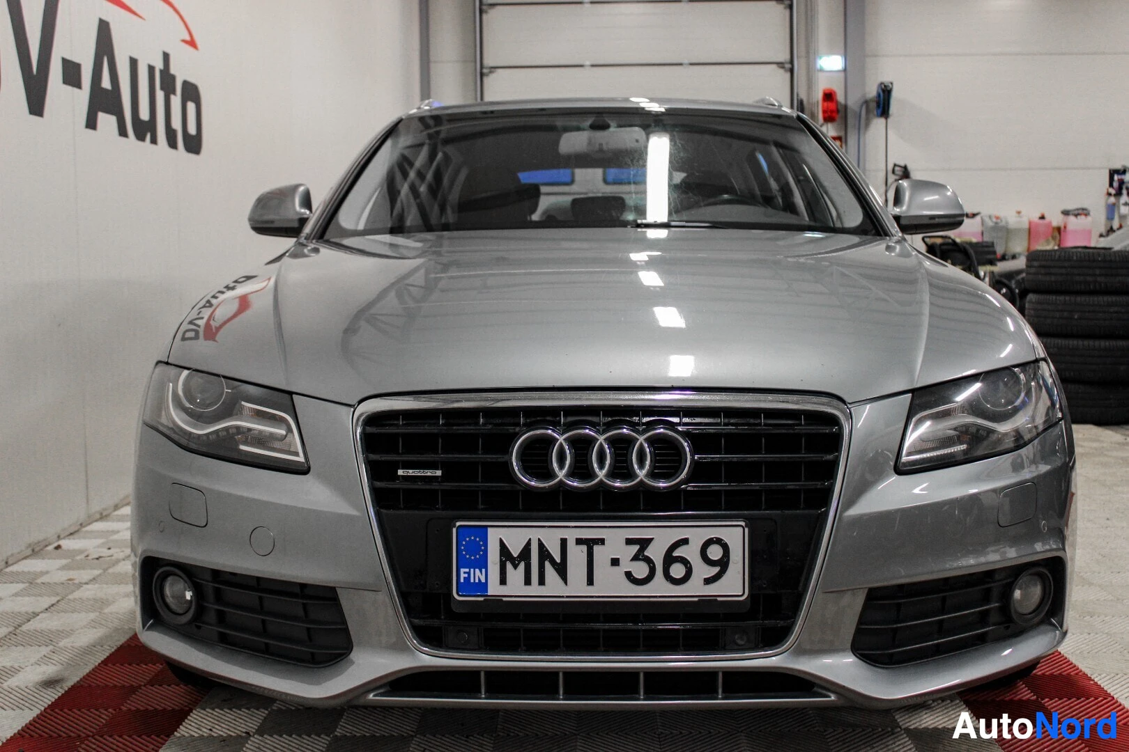 Edestä: Harmaa Audi A4 Avant -farmariauto kuvattuna suoraan edestä autohallissa. Edessä on näkyvillä rekisterikilpi. Valo heijastuu konepellistä ja tuulilasista