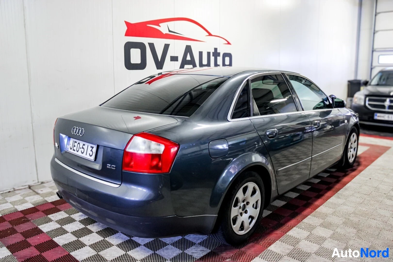 Takaviisto: Tummanharmaa Audi A4 -sedan on pysäköity autonäyttelytilan lattialle, jossa on ruutukuvioinen lattia. Auto on kuvattu takaapäin oikealta puolelta, ja sen rekist