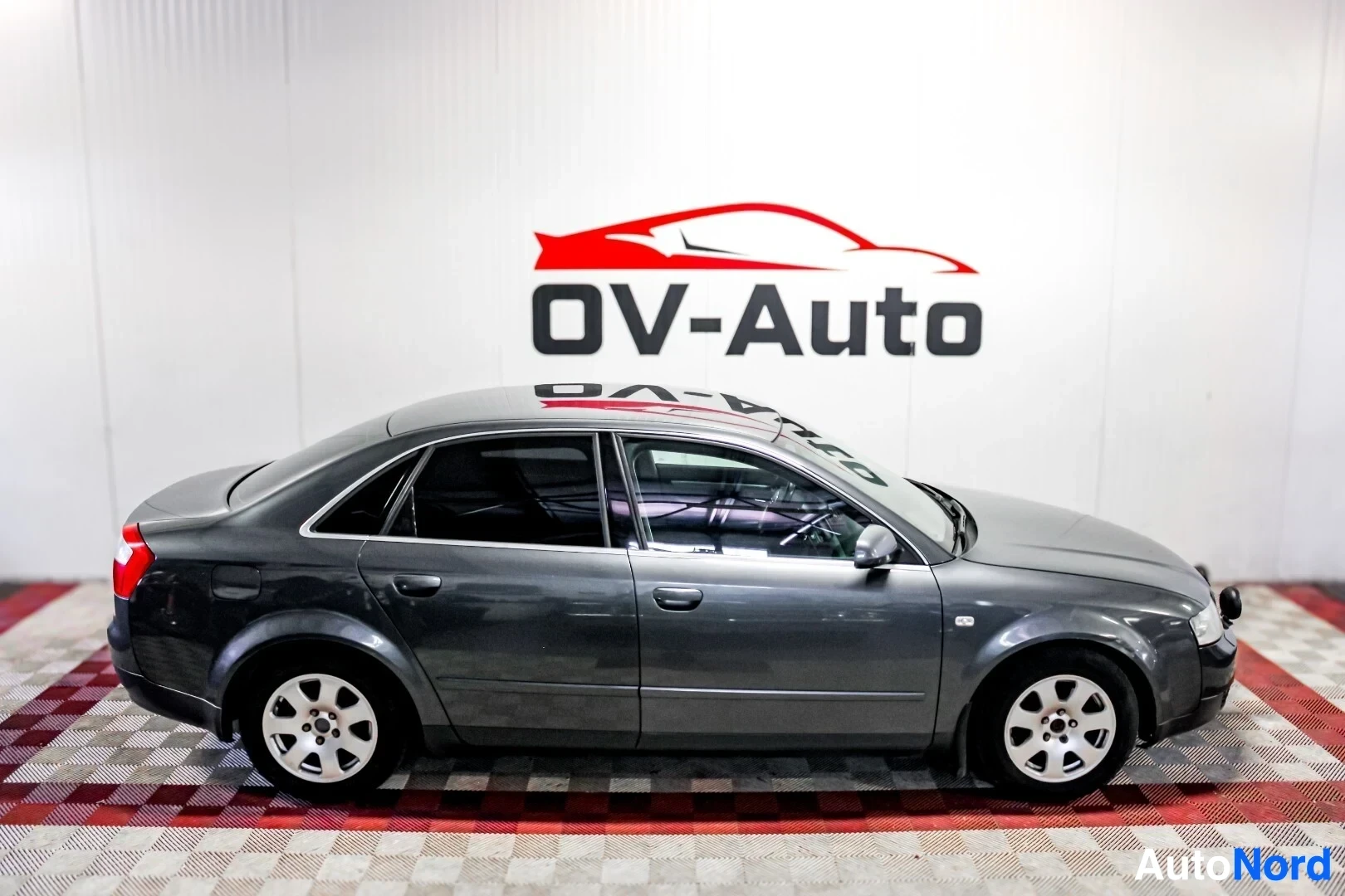 Sivulta: Tummanharmaa Audi A4 -sedan on kuvattu vasemmalta sivulta. Auto seisoo ruutukuvioisella lattialla, ja sen vasen kylki on kokonaan näkyvissä. Valkoiseen seinään