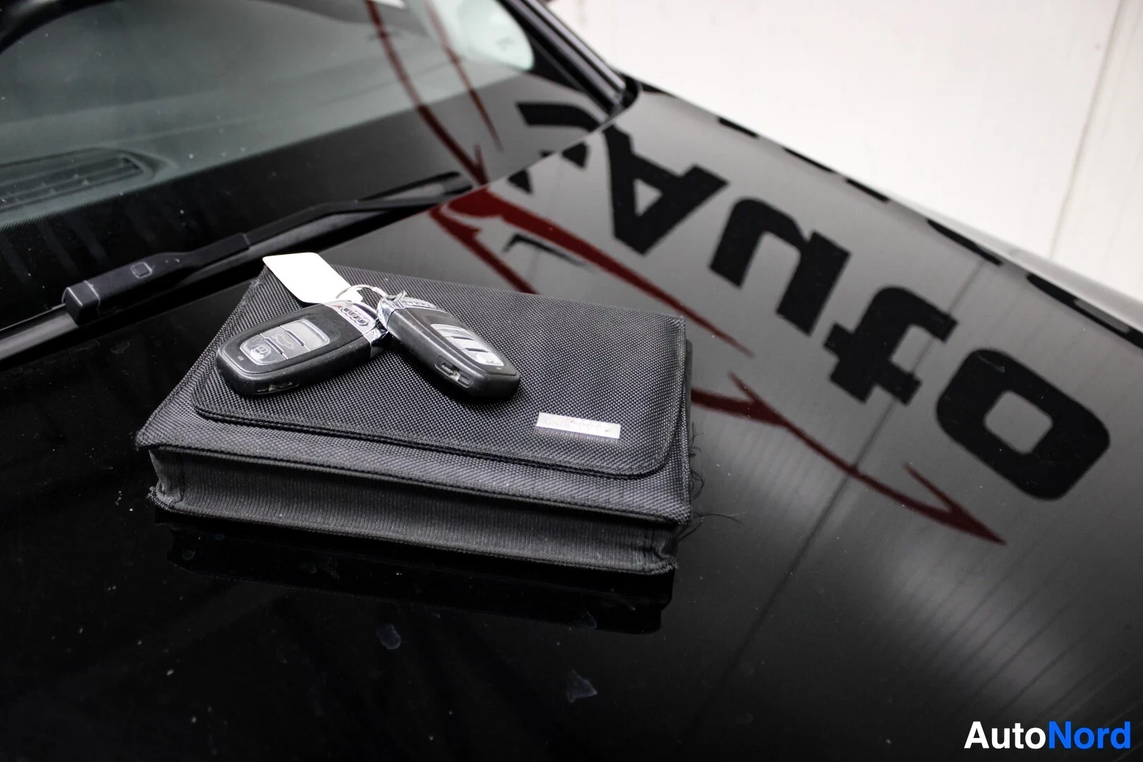 Yksityiskohta: Kaksi auton avainta ja musta ohjekirjalaukku mustan konepellin päällä.
