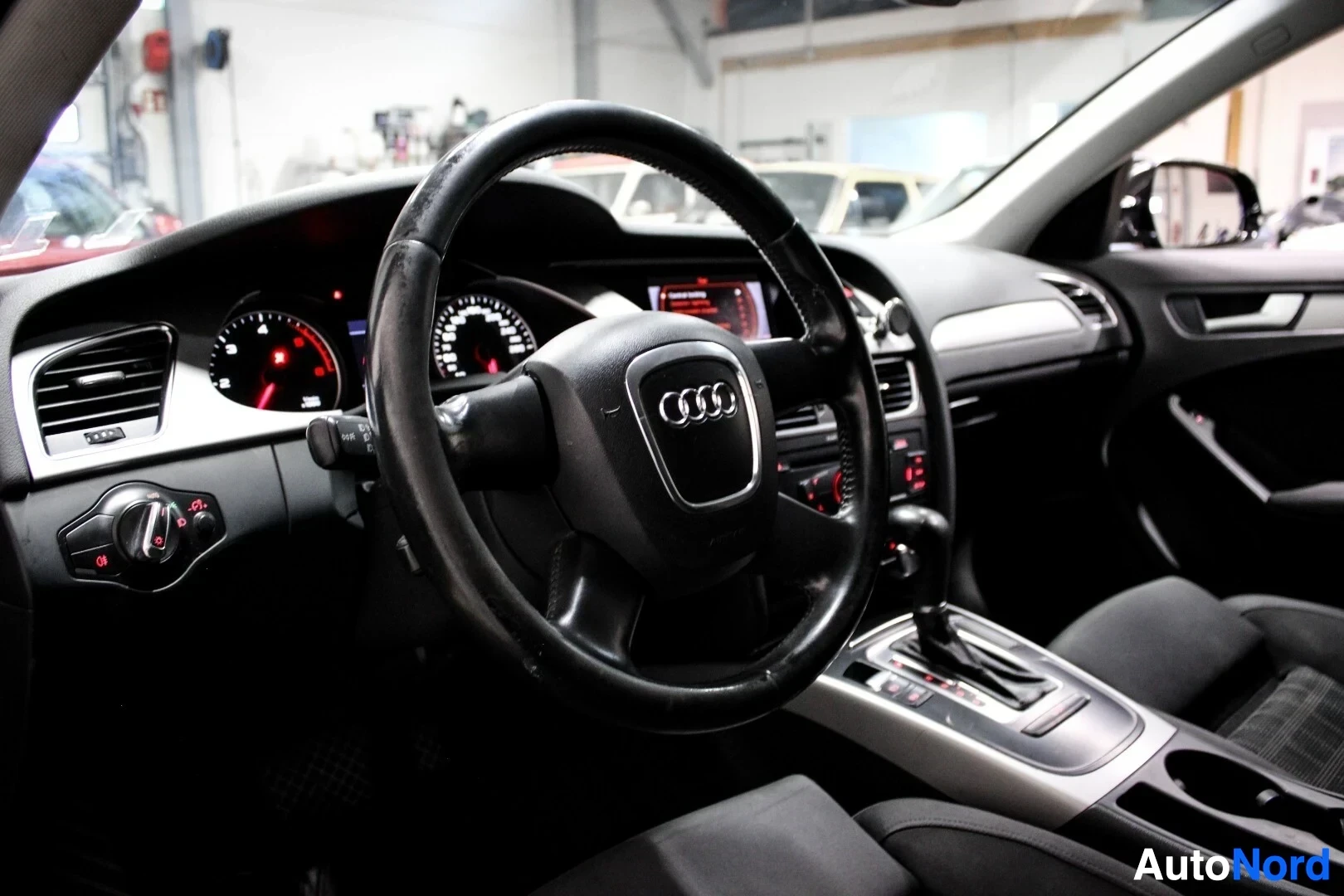 Ohjaamo: Kuvassa on auton ohjaamo, jossa näkyvät ohjauspyörä, mittaristo ja keskikonsolin painikkeet. Ohjauspyörässä on Audi-logo ja mittaristossa valaistut mittarit.
