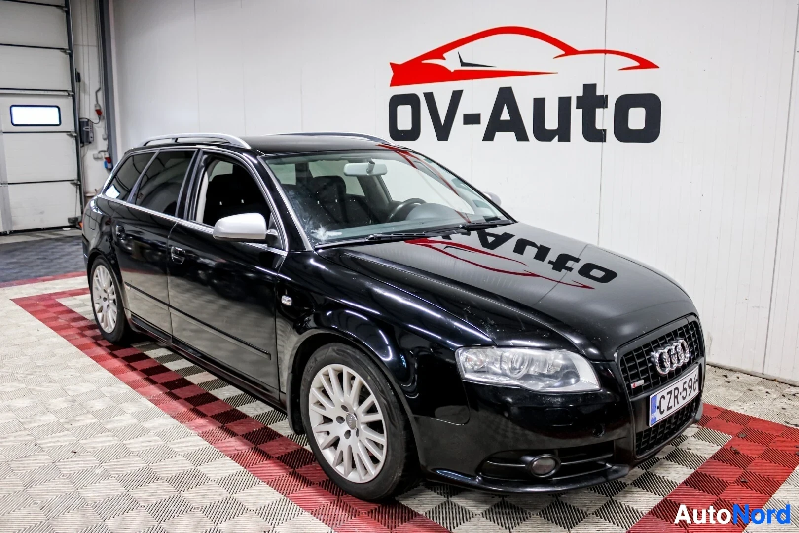 Etuviisto, nokka oikealle: Musta farmariauto, jossa on kattokaiteet ja vaaleat kevytmetallivanteet, seisoo vinosti studiossa. Auton keula on kuvassa oikealla.