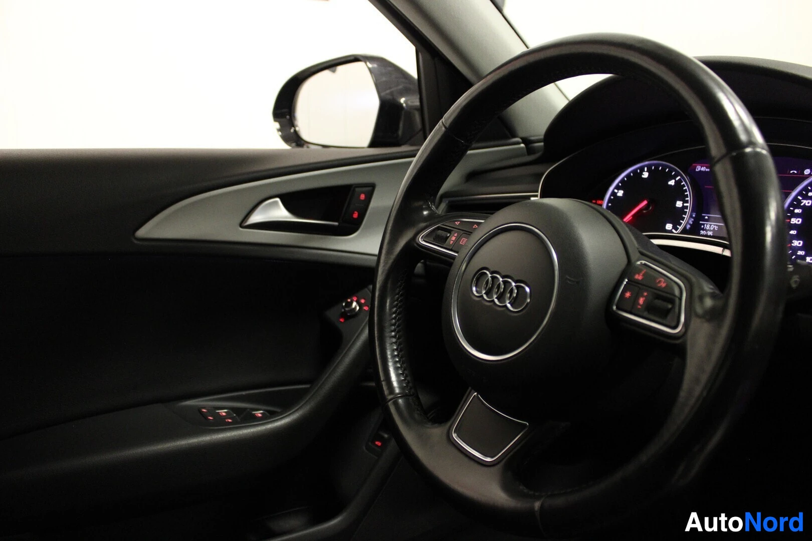 Yksityiskohta: Lähikuva ohjauspyörästä, jossa on hopeinen Audi-logo. Ohjauspyörän oikealla puolella on painikkeita. Myös vasemmanpuoleinen ovipaneeli ja sen ikkunoiden ohjausp