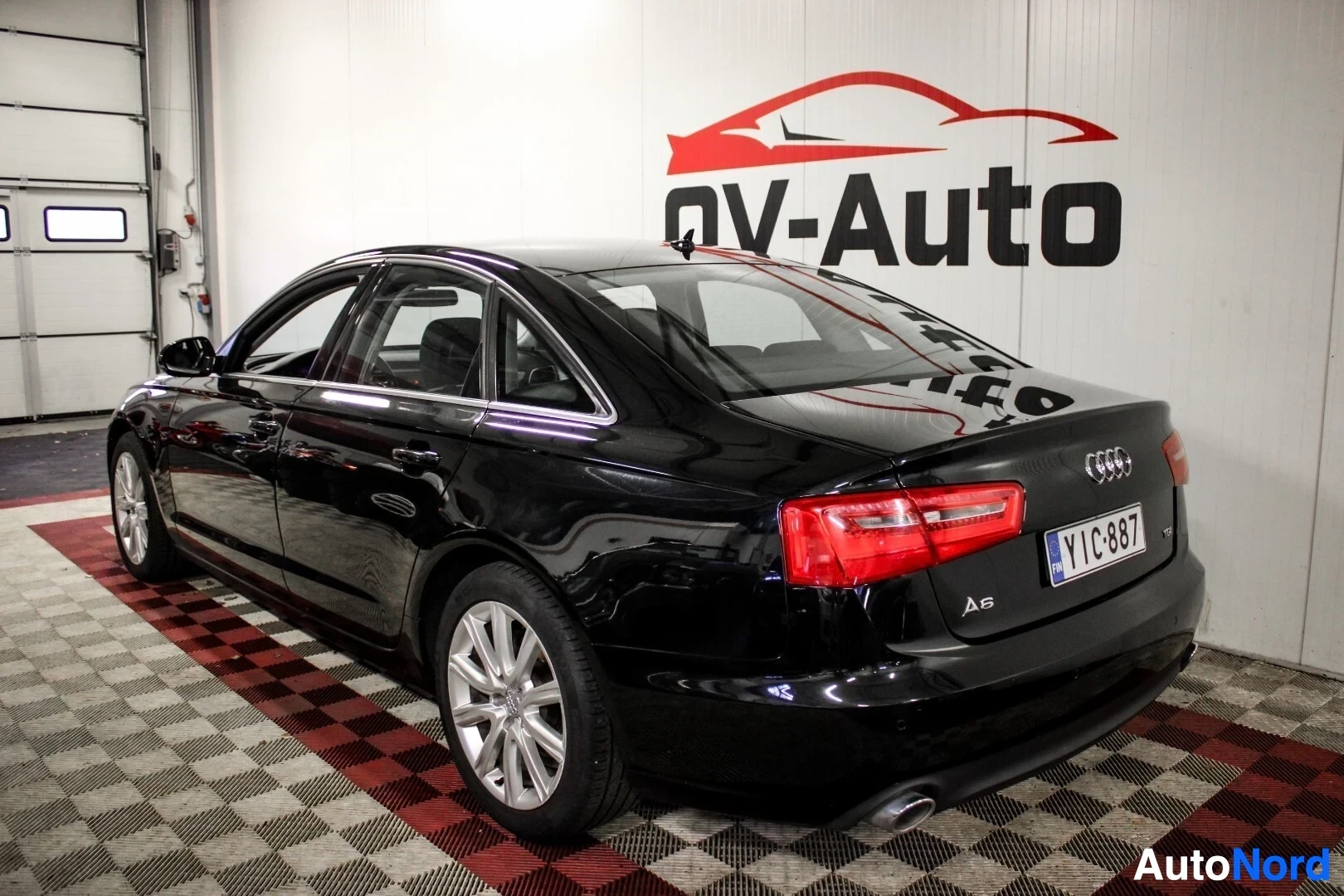 Takaviisto: Musta Audi A6 -henkilöauto takavasemmalta kuvattuna, ajoneuvon rekisterikilvessä on tunnus ja peräkontissa näkyy 'A6'-merkki. Autossa on hopeanväriset k