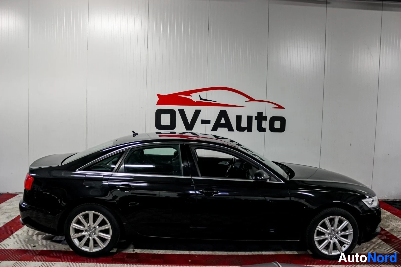 Sivulta: Musta Audi A6 -henkilöauto oikealta sivulta kuvattuna. Autossa on viisipuolaiset hopeanväriset kevytmetallivanteet. Ajoneuvo on pysäköity ruudulliselle lattiall