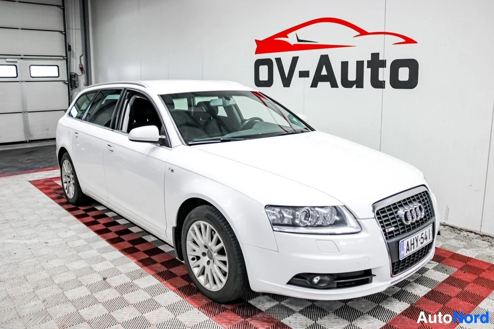 Etuviisto, nokka vasemmalle: Valkoinen Audi A6 farmariauto kuvattu edestä vasemmalta viistosti OV-Auto-logolla varustetussa huoneessa. Autossa on kattokiskot ja monipuolaiset kevyt