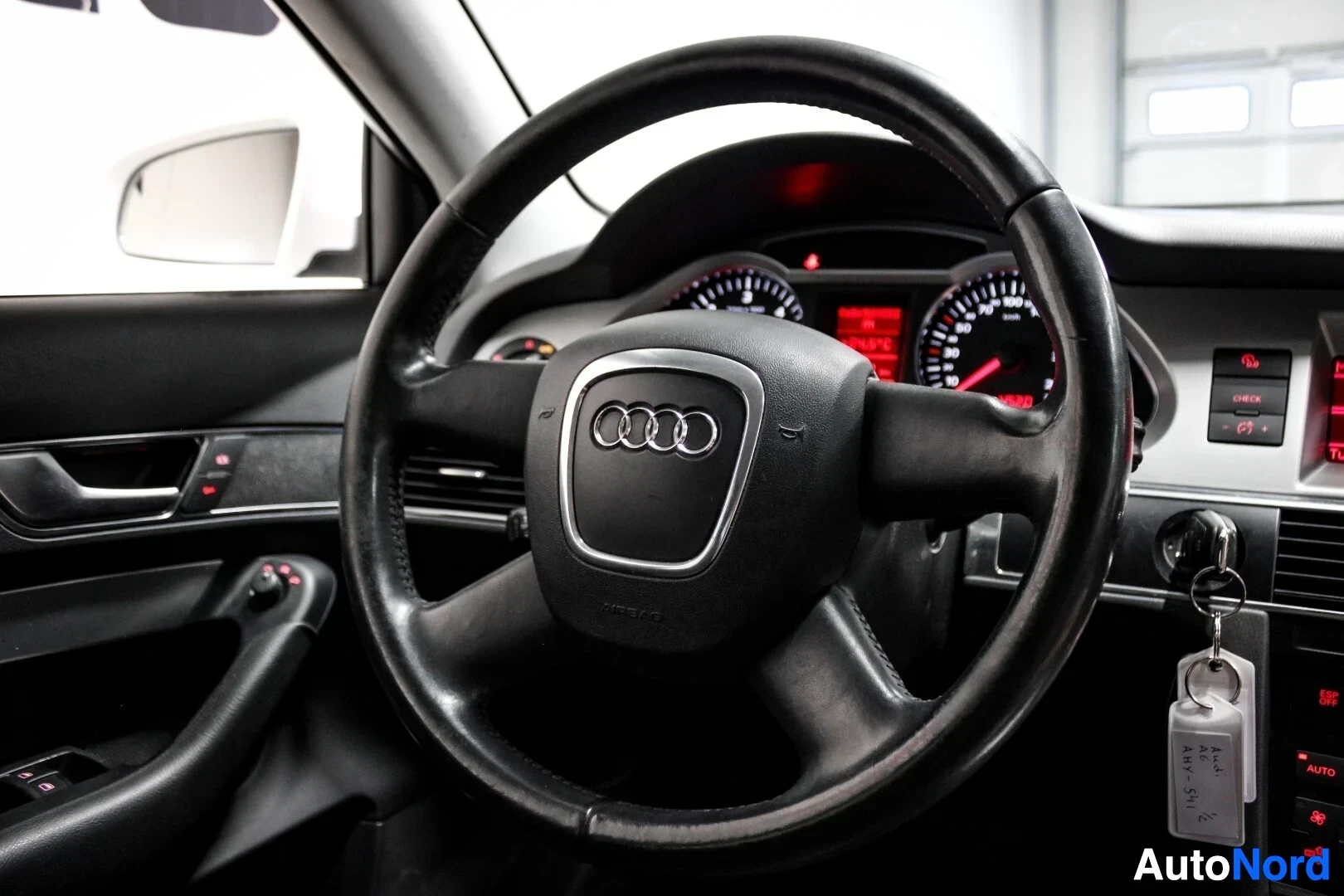 Yksityiskohta: Lähikuva ohjauspyörästä, jossa on Audi-logo keskellä ja painikkeita vasemmalla puolella. Mittariston näytöt ovat valaistuna taustalla.