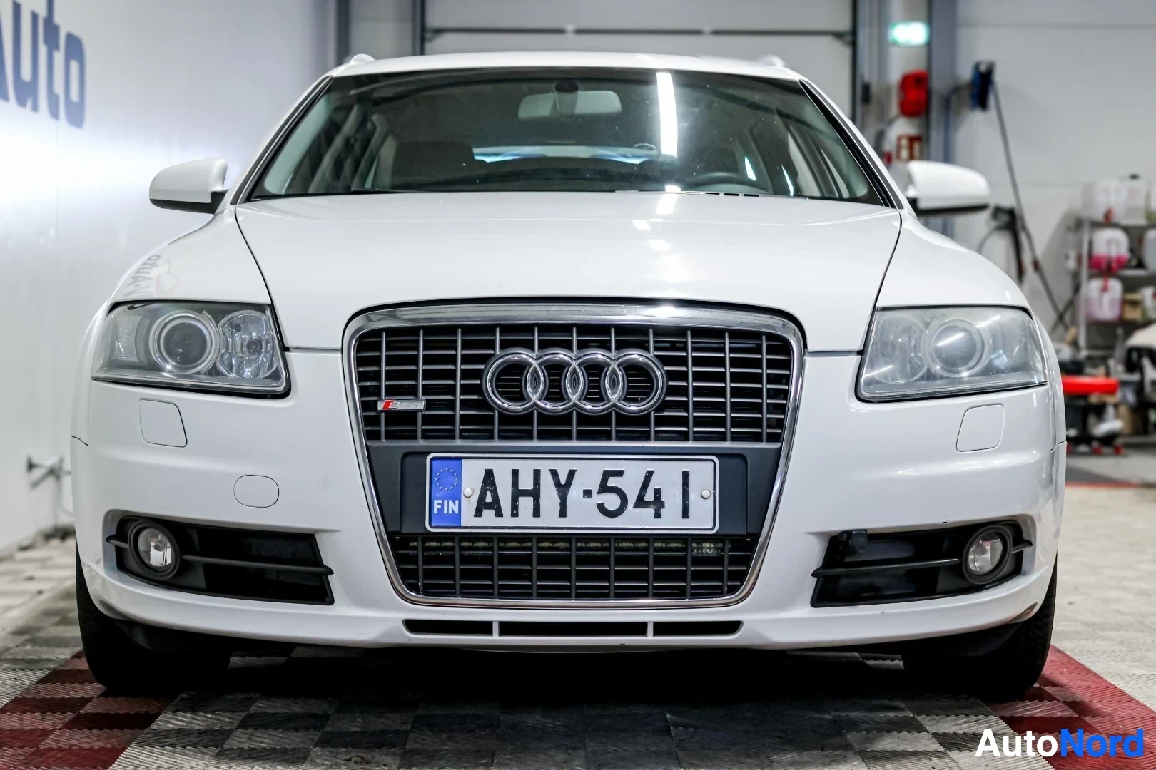 Edestä: Valkoinen Audi A6:n keula. Autossa on neljä rengasta logona etusäleikössä ja rekisterikilpi "FIN "