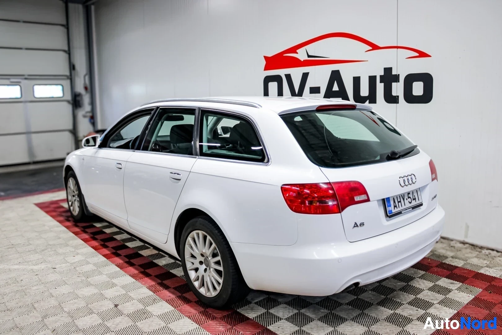 Takaviisto: Valkoinen Audi A6 farmariauto kuvattu takaa vasemmalta viistosti OV-Auto-logolla varustetussa huoneessa. Autossa on kattokiskot ja rekisterikilpi ""