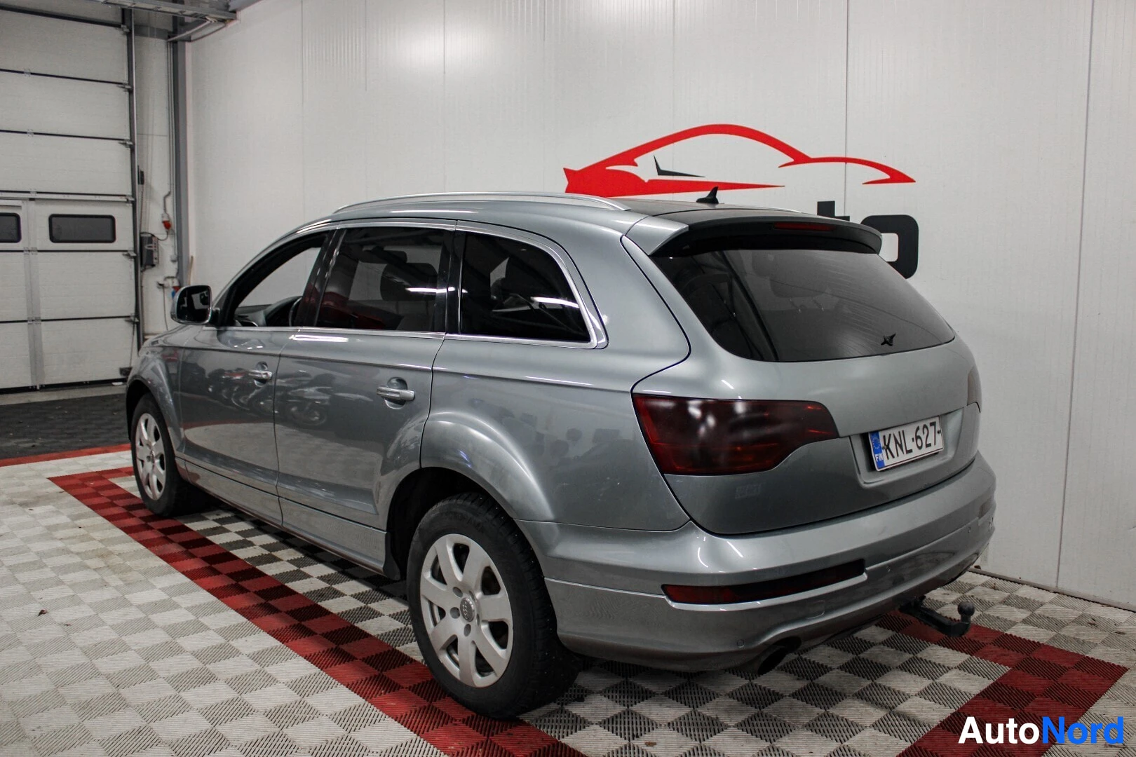 Takaviisto: Harmaa Audi Q7 -maastoauto pysäköitynä sisätiloihin vasemmassa takaviistokuvassa. Ajoneuvossa on kattokaiteet, alumiinivanteet ja vetokoukku.