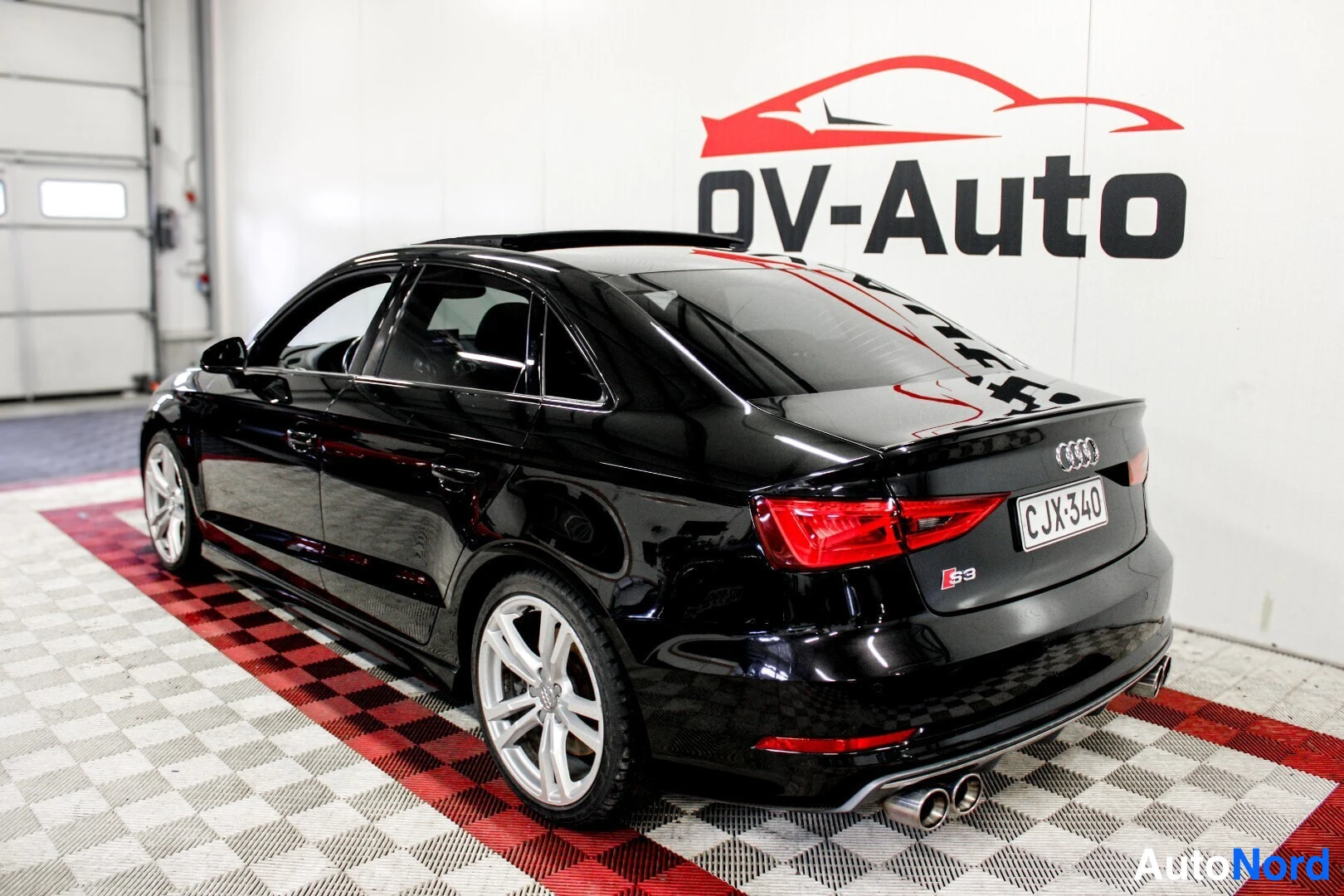 Takaviisto: Musta Audi S3 -henkilöauto takavasemmalta kuvattuna. Autossa on kattoluukku ja vaaleat kevytmetallivanteet. Taustalla näkyy seinä, jossa on "OV-Auto"-logo ja pu