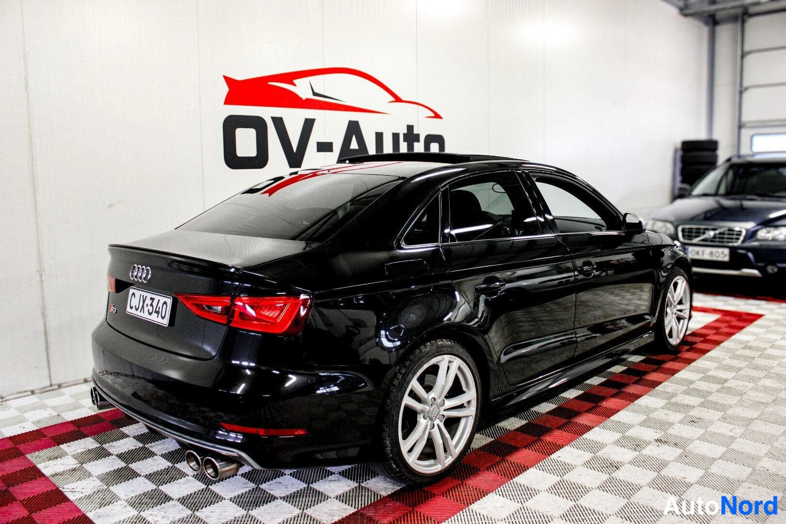 Takaviisto: Musta Audi S3 -henkilöauto takavasemmalta kuvattuna. Autossa on kattoluukku ja vaaleat kevytmetallivanteet. Taustalla näkyy seinä, jossa on "OV-Auto"-logo ja pu