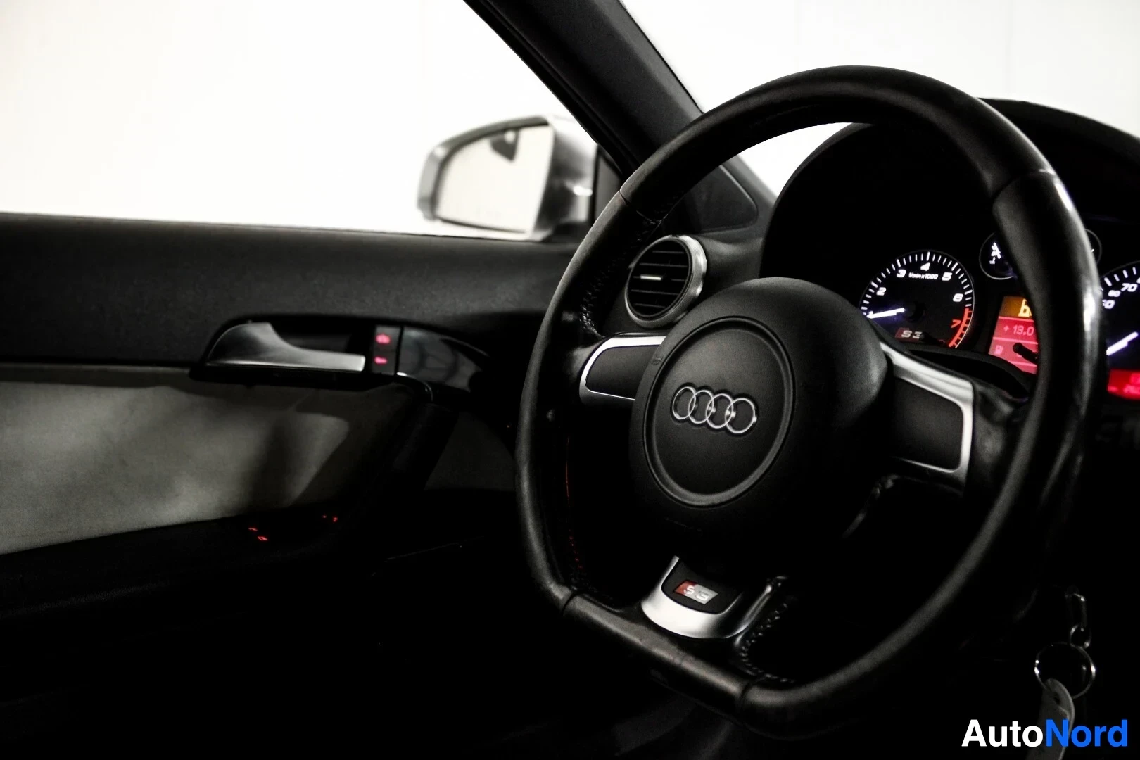 Ohjaamo: Lähikuva auton ohjauspyörästä kuljettajan puolelta. Ohjauspyörässä on Audi-logo ja 'S3' -merkki. Ovipaneelissa näkyy ovenkahva ja peilin säätimet.