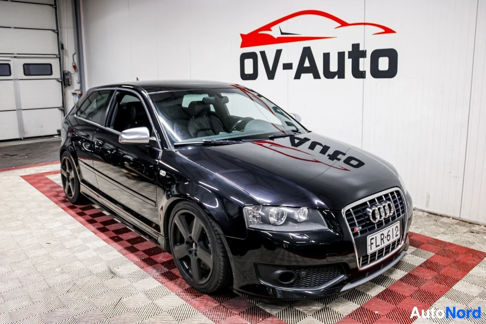 Etuviisto, nokka oikealle: Musta kolmiovinen Audi S3, jossa on tummat kevytmetallivanteet ja hopeiset sivupeilit, pysäköitynä vinosti OV-Auton logolla varustetun seinän eteen.