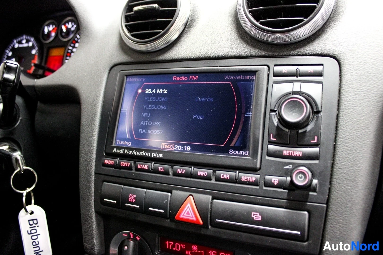 Yksityiskohta: Lähikuva auton keskikonsolista, näyttäen radionäytön, jossa on näkyvissä FM-radioasemia, ja ilmastoinnin hallintalaitteet. Näytöllä on teksti "Audi Navigation p