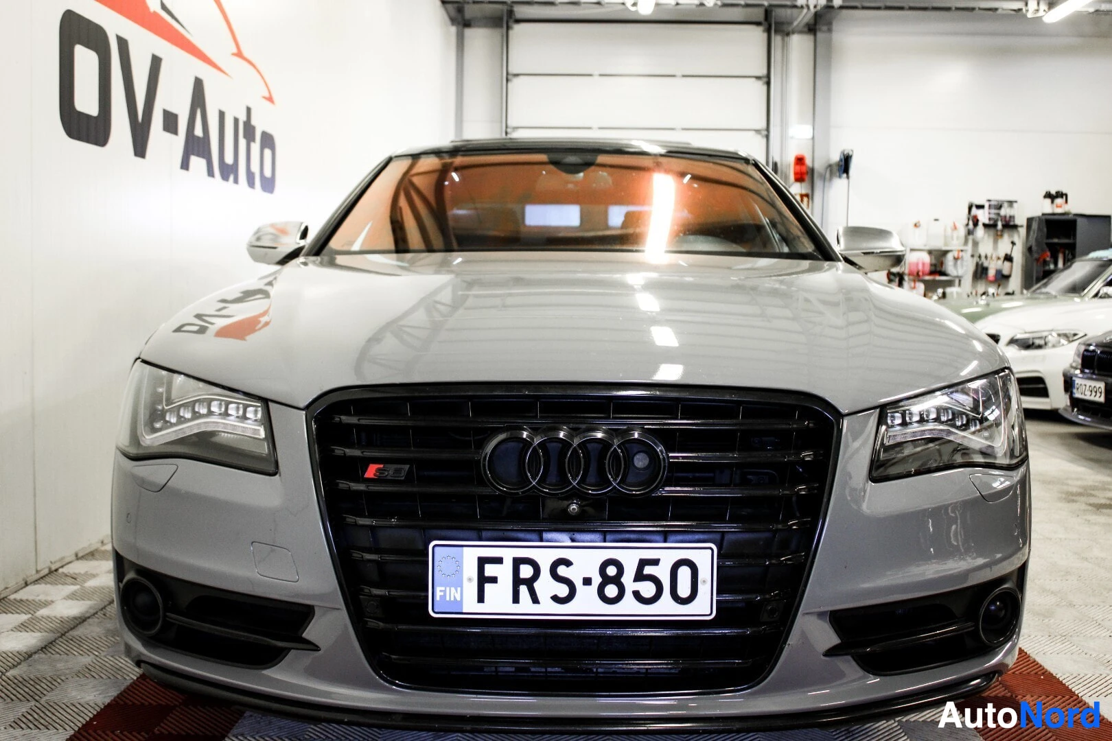 Edestä: Harmaa Audi S8 edestä kuvattuna. Auton etupuskurissa on musta ritiläsäleikkö ja rekisterikilpi. Taustalla on valkoinen seinä, jossa lukee OV-Auto. Etuvalot ovat