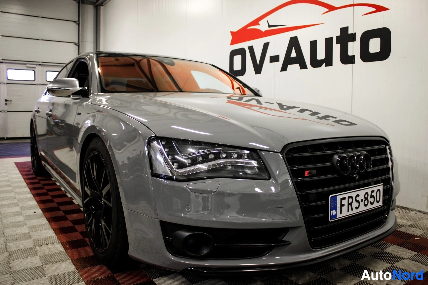 Etuviisto, nokka vasemmalle: Harmaa Audi S8 -henkilöauto pysäköitynä halliin, sen vasen etukulma näkyvissä. Autossa on mustat monipuolaiset vanteet ja etusäleikössä on -reki