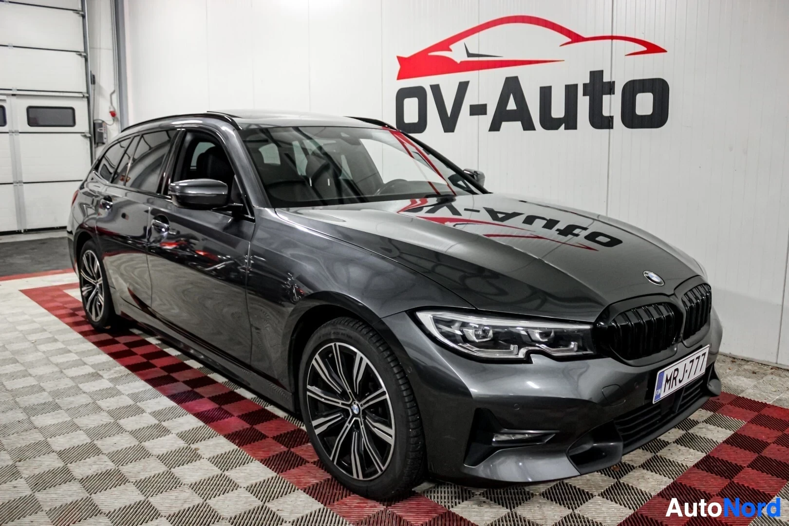 Etuviisto, nokka vasemmalle: Harmaa BMW 3-sarjan farmari pysäköitynä myyntihallissa, näkyy etuoikealta.