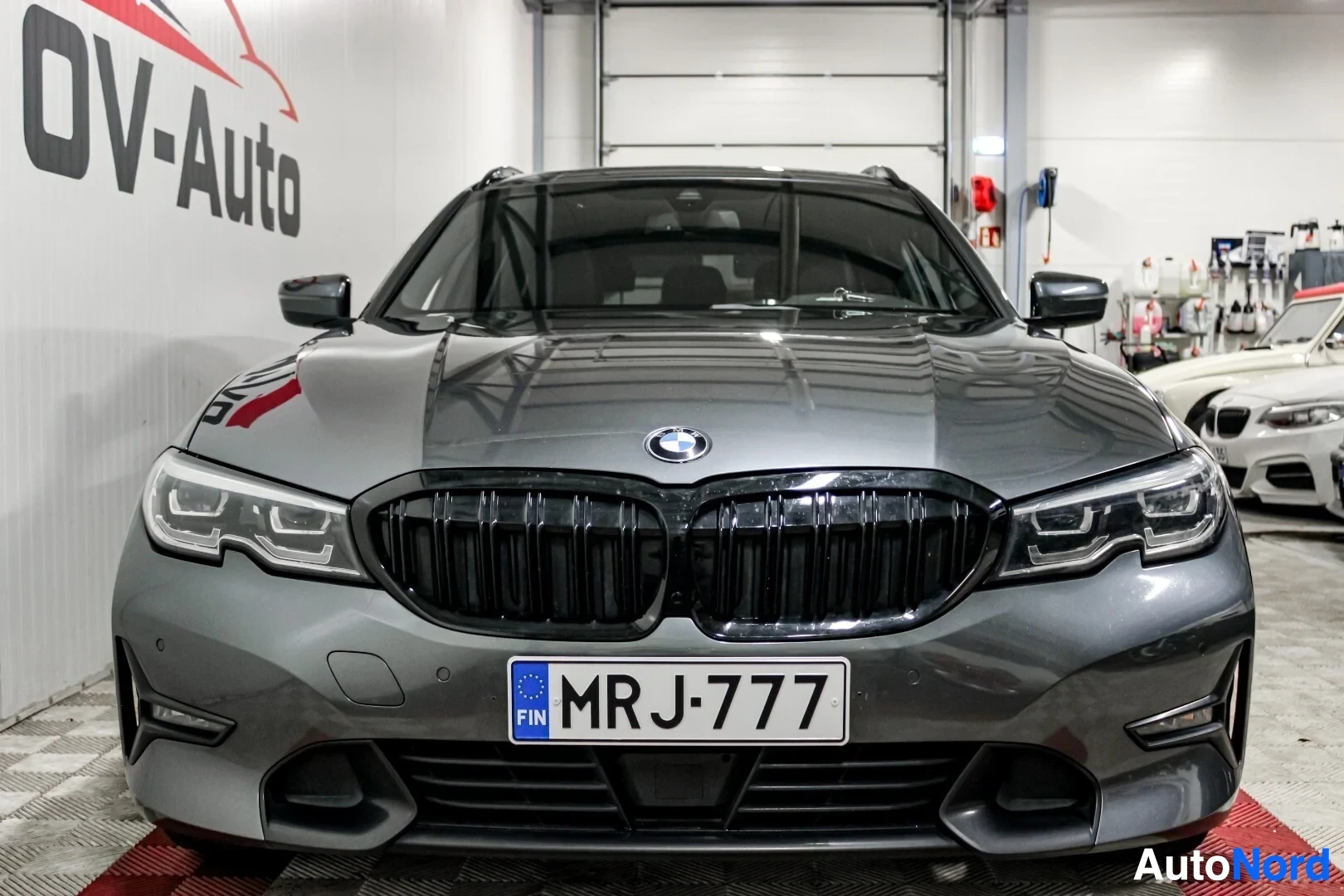 Edestä: Harmaa BMW 3-sarjan farmari myyntihallissa, näkyy suoraan edestä.