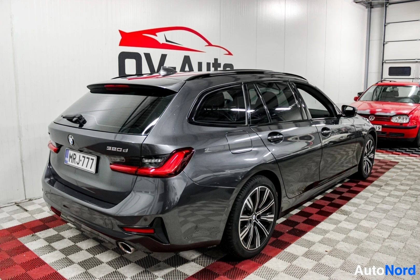 Takaviisto: Tummanharmaa farmariauto kuvattuna taka-viistosta vasemmalta puolelta sisätiloissa. Autossa on kattokaiteet ja monipuolaiset kevytmetallivanteet. Takaluukussa n