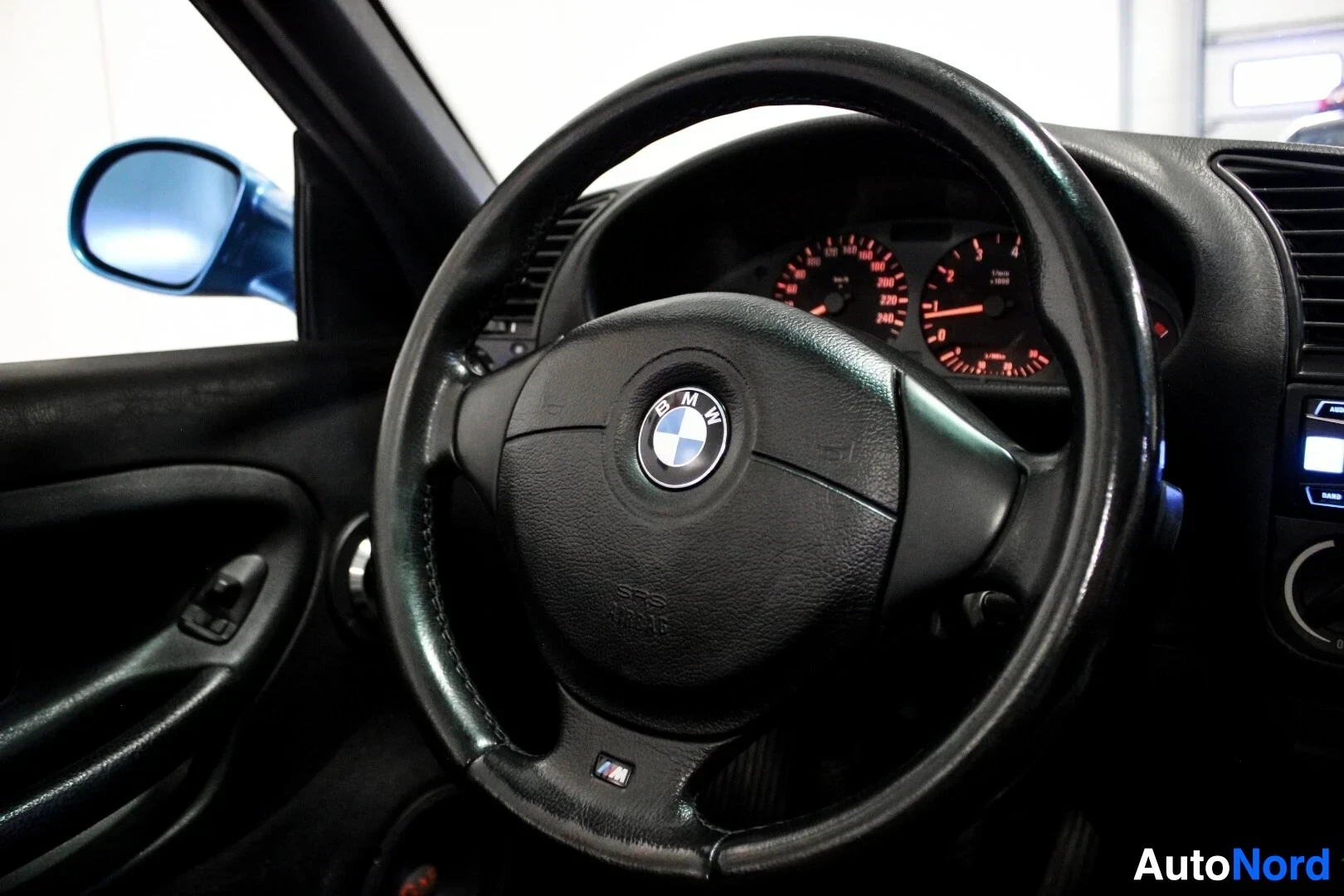 Ohjaamo: Lähikuva ohjauspyörästä ja mittaristosta kuljettajan puolelta. Ohjauspyörässä on BMW-logo ja alareunassa M-logo. Taustalla näkyy vasen sivupeili.