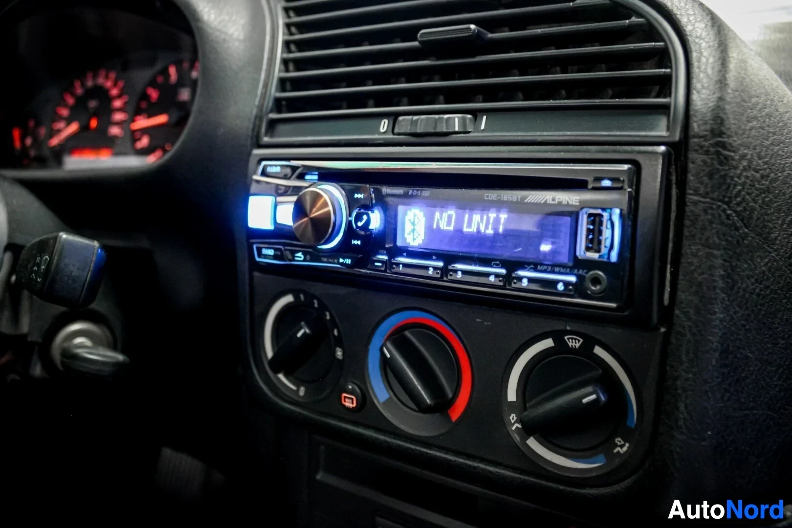 Ohjaamo: Lähikuva auton keskikonsolista, jossa on Alpine-merkkinen radio ja ilmastoinnin säätimet. Radion näytöllä lukee "NO UNIT" ja Bluetooth-kuvake.