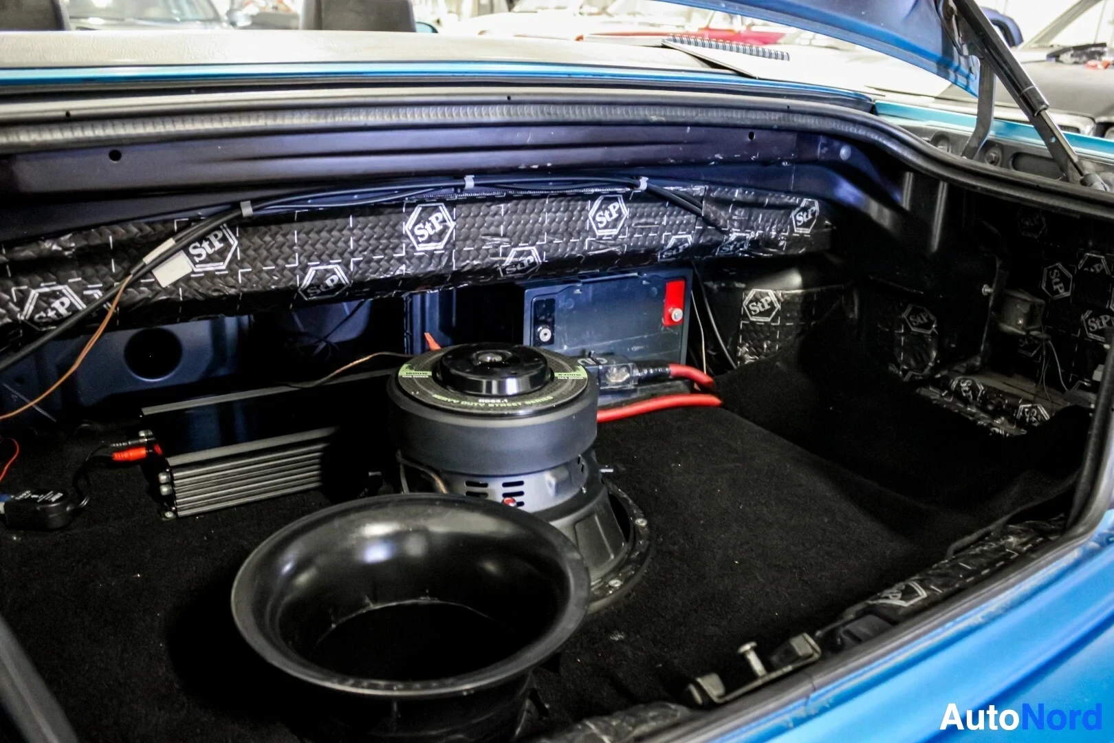 Tavaratila: Kuva auton tavaratilasta, jossa on asennettuna subwoofer, vahvistin, ylimääräinen akku ja äänenvaimennusmateriaaleja.