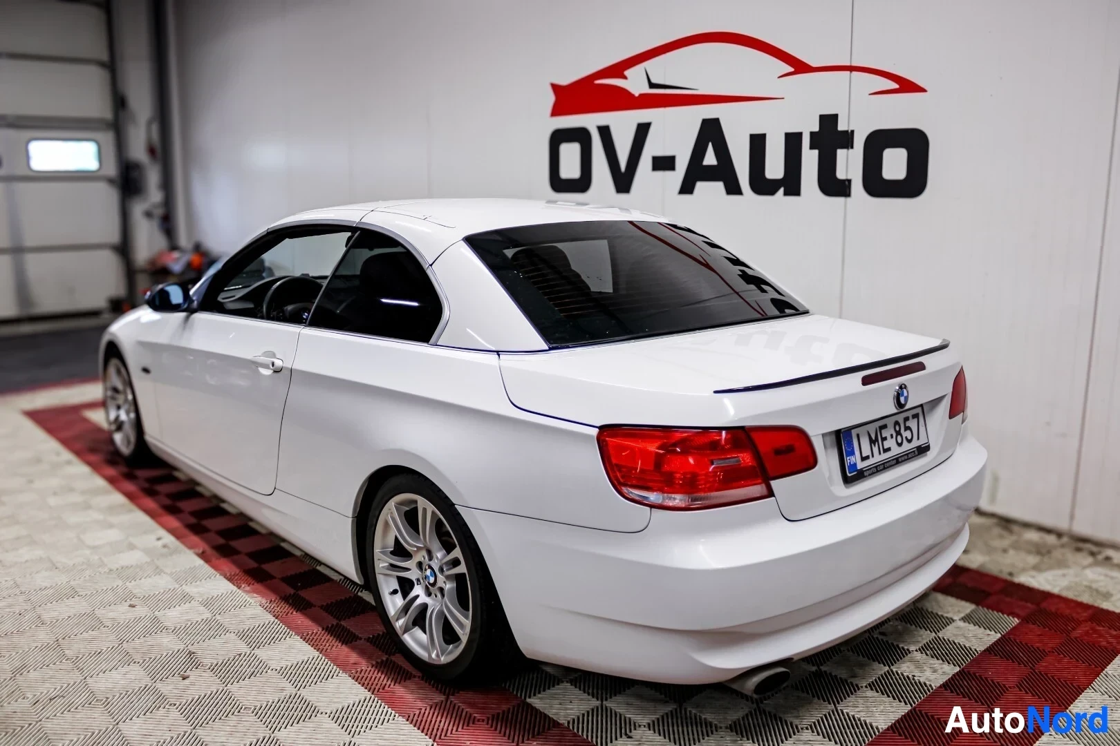 Takaviisto: Valkoinen BMW 3-sarjan avoauto takaa ja vasemmalta sivulta kuvattuna autoliikkeen tiloissa, takana rekisterikilpi