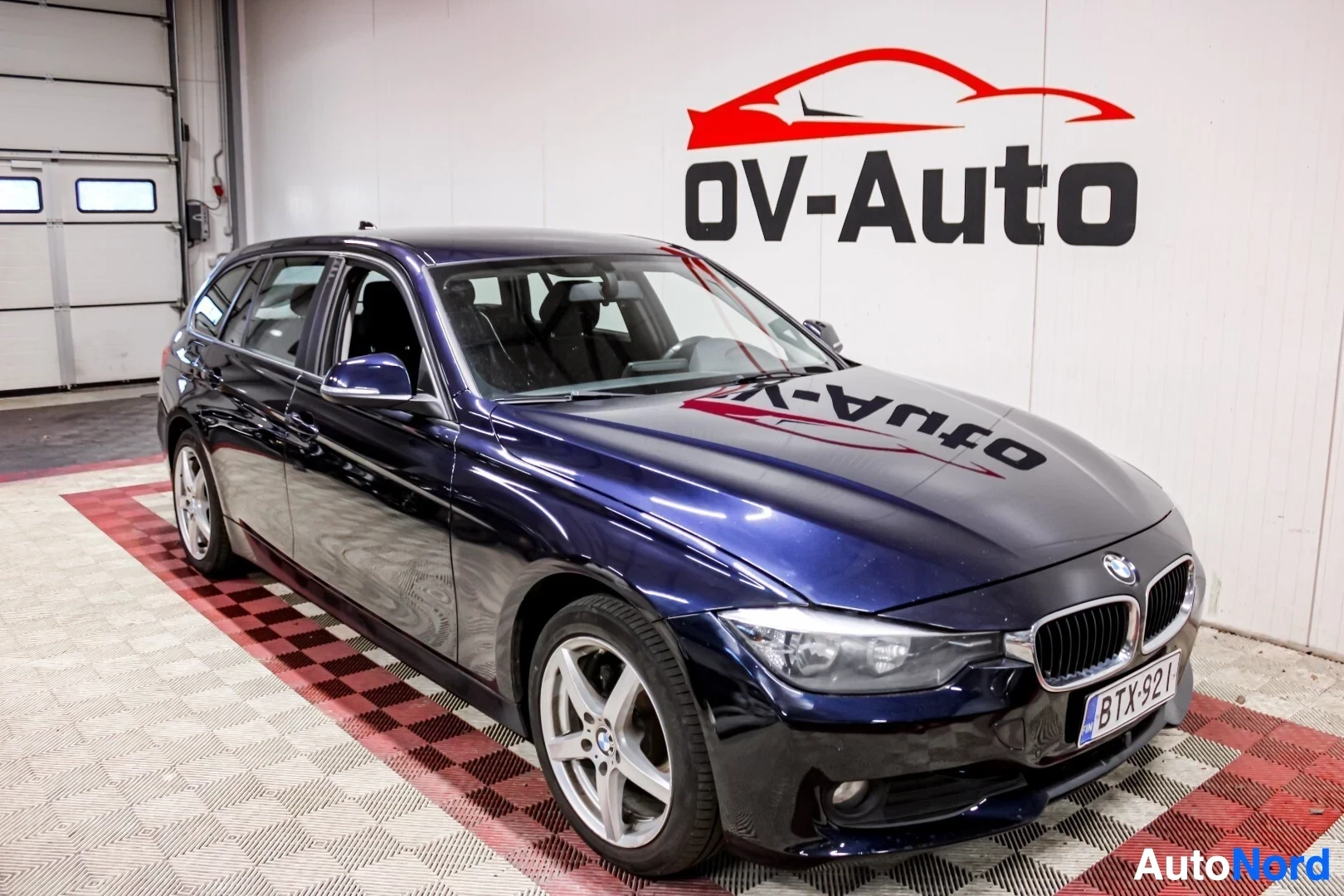 Etuviisto, nokka oikealle: Sivutakakulmasta kuvattu tummansininen BMW farmariauto OV-Auton logolla varustetun seinän edessä. Ajoneuvossa on hopeiset kevytmetallivanteet ja kattokis