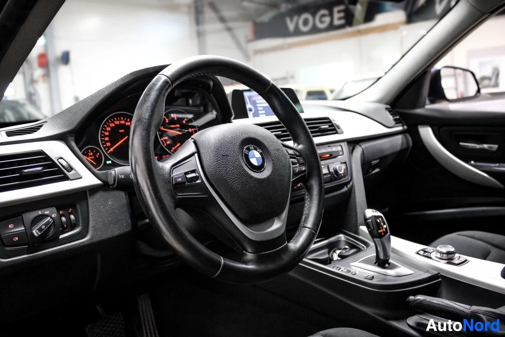 Ohjaamo: Kuvassa on auton ohjaamo, jossa näkyy ohjauspyörä, mittaristo ja keskikonsoli. Ohjauspyörässä on BMW-logo ja monitoiminäppäimet. Mittaristossa on oranssi valais